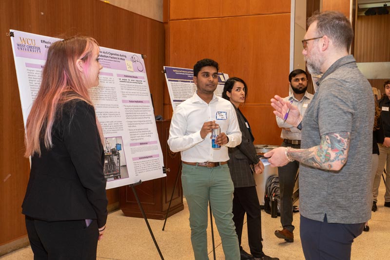 Image from a past years Research and Creative Activity Day - a group of people stand around chatting in front of poster board