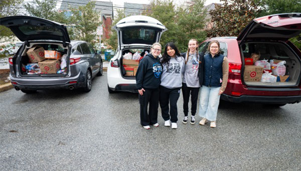 Photo of 4 students standing in front of cars with their trunks full of food items