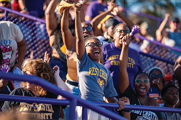 A picture of the crowd in the bleachers but a young girl is the focus. She is smiling and raising her hand in a celebratory manner.
