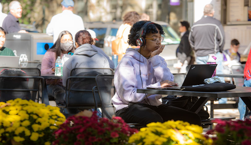 A student sits outside on a picnic bench wearing headphones and using her laptop. Yellow and purple flowers are in the foreground.