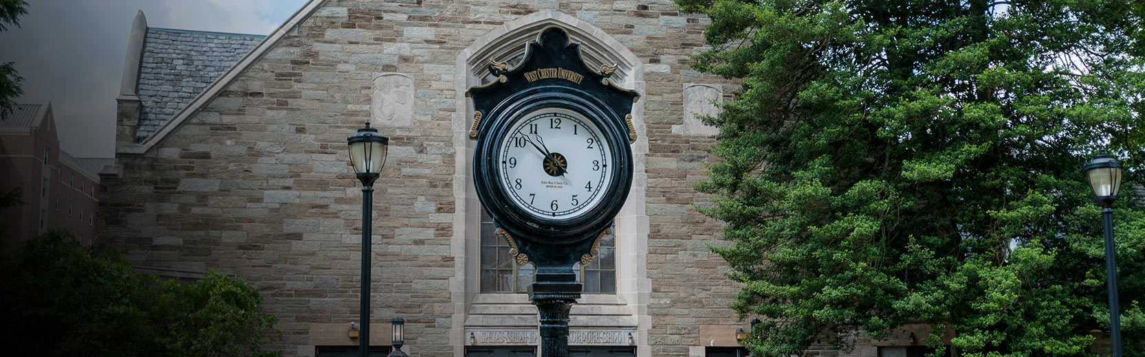 Large freestanding campus clock in front of a stone university building, surrounded by trees and lampposts.